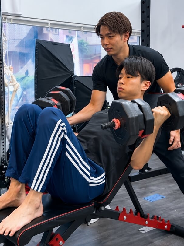 トレーナーが見守る中、男性がベンチに座ってダンベルを持ち上げるトレーニングを行っている様子