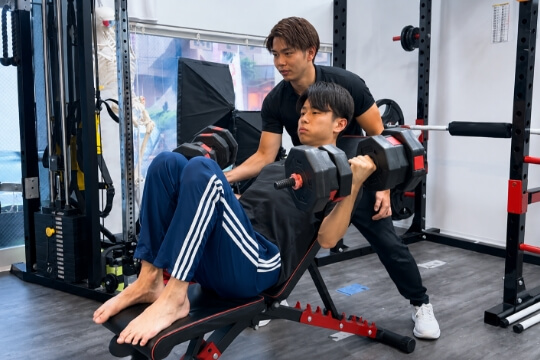 トレーナーが見守る中、男性がベンチに座ってダンベルを使ったトレーニングを行っている様子