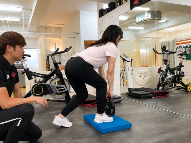 トレーナーが見守る中、女性がバランスパッドの上でウエイトを持ってトレーニングを行っている様子