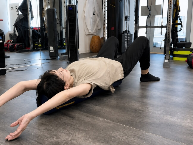 女性がマットの上で仰向けになり、腕を広げて体を反らせるストレッチをしている様子