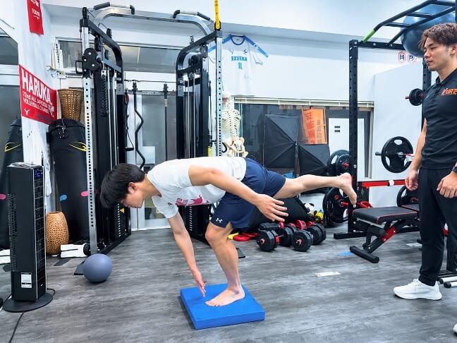トレーナーが見守る中、男性がバランスパッドの上で前傾姿勢のトレーニングを行っている様子